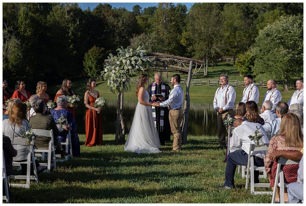 Bride and groom holding hands at ceremony saying “I do” outside with green trees in the background