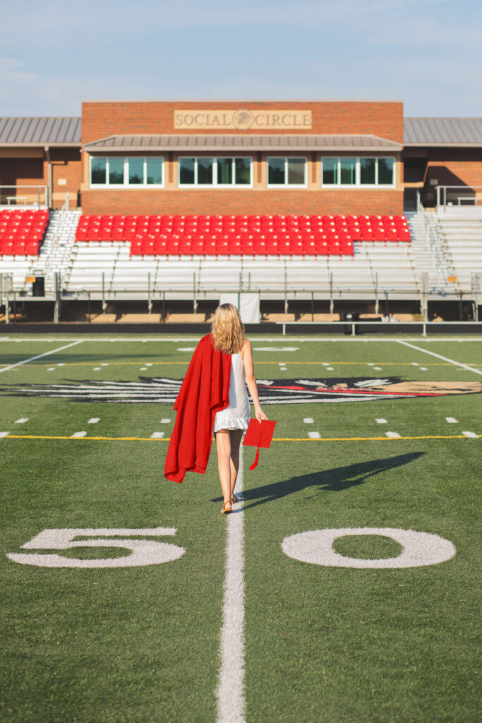 A young woman wearing a red graduation gown and holding a diploma walks down the 50-yard line of a football field with bleachers and a building labeled "SOCIAL CIRCLE" in the background.